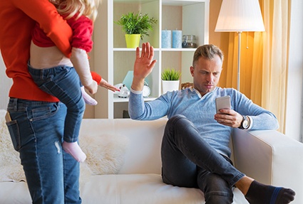 Mann sitzt auf Sofa, vor ihm Frau mit schreiendem Kind, er währt mit der Hand ab, und schaut auf sein Smartphone
